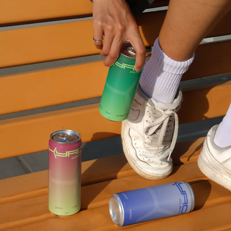 A person sitting on a wooden bench holds a green Kyro can, with pink and blue Kyro cans nearby.